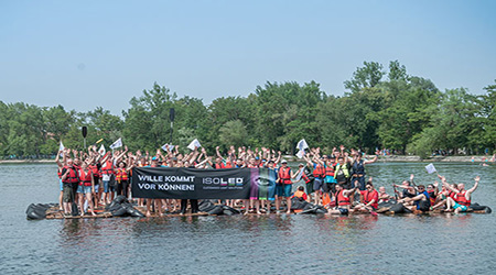 Große Gruppe von Menschen mit Schwimmwesten auf einem selbstgebauten Floß auf einem See, hält ein Banner mit der Aufschrift Wille kommt vor Können! ISOLED, umgeben von Bäumen.
