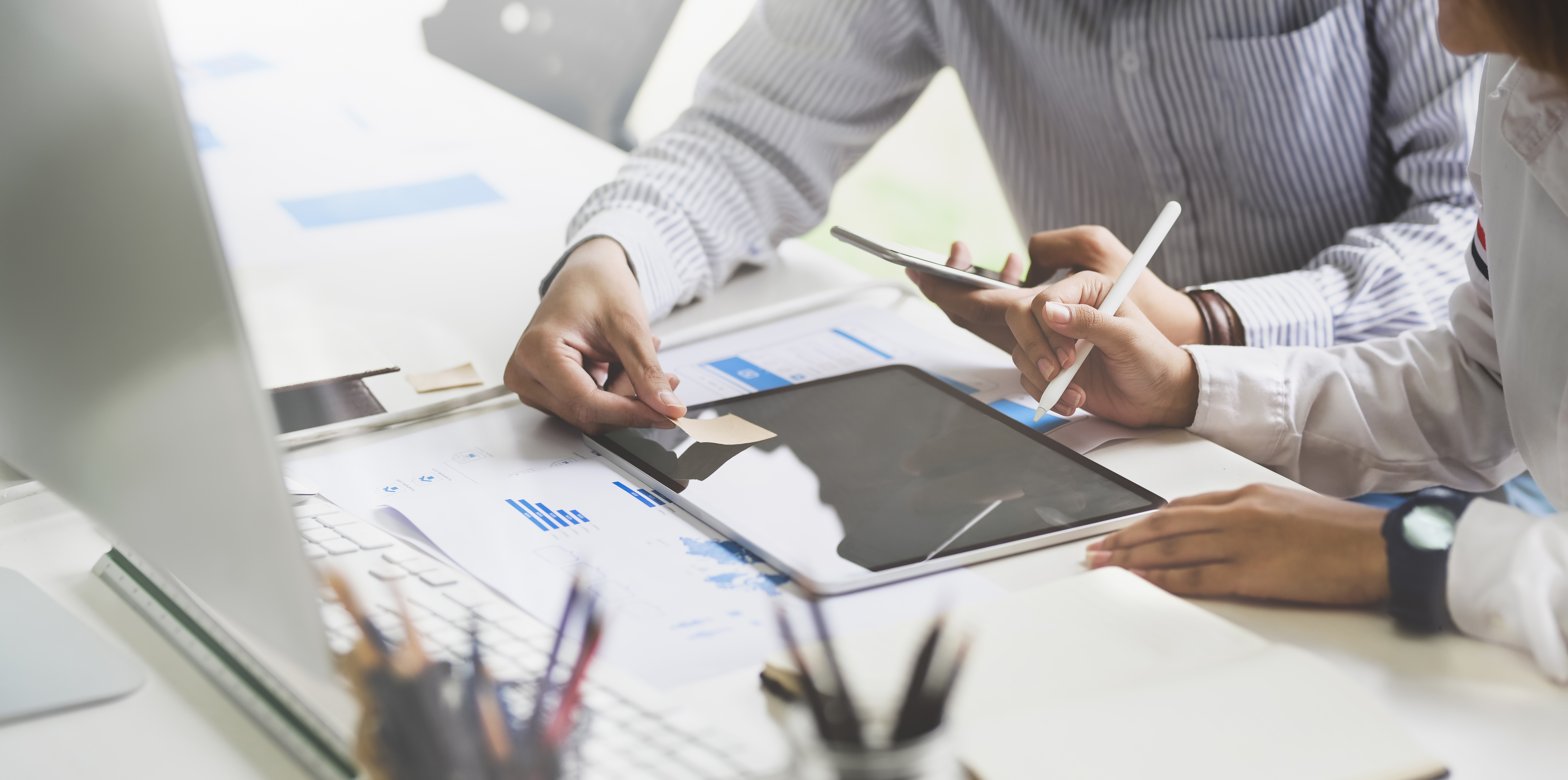 Tablet &ndash; Zwei Personen diskutieren und arbeiten mit einem Tablet am Schreibtisch, umgeben von Dokumenten und B&uuml;romaterialien in einer modernen B&uuml;roumgebung.