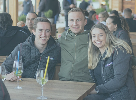 Drei junge Menschen sitzen lächelnd an einem Holztisch mit Getränken in Gläsern mit bunten Strohhalmen, in einem belebten Restaurant oder Café, tragen ISOLED-Jacken.