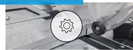 Eine Kreiss&auml;ge schneidet durch ein Metallst&uuml;ck. Ein Zahnradsymbol ist dar&uuml;bergelegt. Der Kontext zeigt eine Werkstattumgebung mit Metallarbeitsfl&auml;che und einer transparenten Schutzabdeckung.